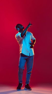 Tam boy gülümseyen Afro-Amerikan bir adam akıllı gündelik tarzda kulaklıkla müzik dinliyor neon ışıkta kırmızı arka planda. İnsan duyguları kavramı yüz ifadesi, moda