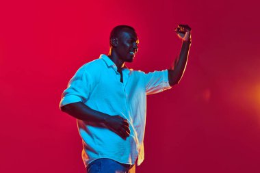 Beyaz gömlekli genç Afro-Amerikan bir adamın portresi, neon ışıkta kırmızı arka planda başarı ve büyümeyi ifade ediyor. İnsan duyguları kavramı yüz ifadesi, moda