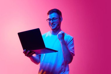 Gözlüklü ve beyaz tişörtlü genç adam dizüstü bilgisayarı ve el kol hareketleriyle pembe arka plandaki neon ışığa karşı başarısını ifade ediyor. İnsan duyguları, yüz ifadesi, yaşam tarzı kavramı