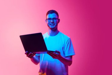 Gündelik beyaz gömlekli, gözlüklü pembe arka planda dizüstü bilgisayarla duran genç bir adam portresi. İşçi. İnsan duyguları kavramı, yüz ifadesi, yaşam tarzı, aletler.