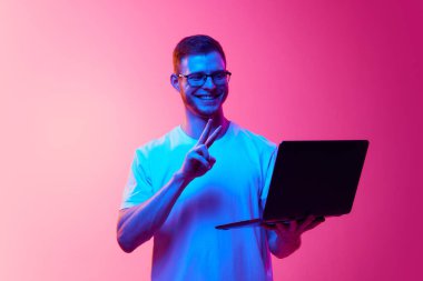 Gözlüklü, beyaz tişörtlü, dizüstü bilgisayarı olan gülümseyen genç bir adam ve pembe arka planda arkadaşlarıyla online video görüşmesi yapan neon ışıklı bir adam. İnsan duyguları, yüz ifadesi, yaşam tarzı kavramı