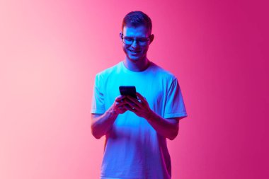 Gözlüklü ve beyaz tişörtlü gülümseyen genç adam iletişim için cep telefonu kullanıyor, mesaj yazıyor, pembe arka planda mesaj yazıyor. İnsan duyguları, yüz ifadesi, yaşam tarzı kavramı