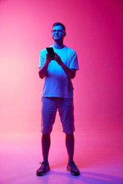Gözlüklü ve sıradan giyinen genç bir adamın pembe arka planda pembe ışıkta iletişim kurmak için cep telefonunu kullanışının tam boy görüntüsü. İnsan duyguları, yüz ifadesi, yaşam tarzı kavramı