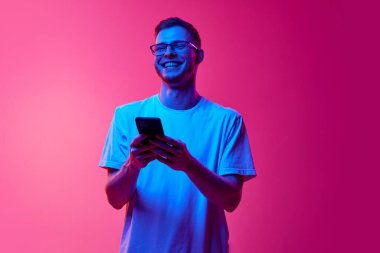 Gözlüklü ve günlük kıyafetlerle gülen genç bir adam cep telefonunu neon ışıkta pembe arka planda iletişim kurmak için kullanıyor. İnsan duyguları, yüz ifadesi, yaşam tarzı kavramı