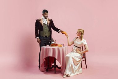 Elegant man and woman in period costumes having breakfast, man giving croissant to attractive woman against pink studio background. Concept of history, retro and vintage, comparison of eras clipart