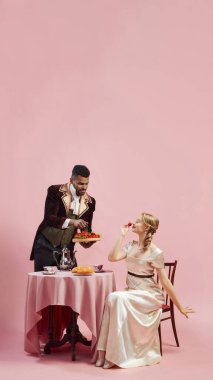 Man in traditional attire presents plate of berries to lady against pink studio background. Gentleman and lady having breakfast. Concept of history, retro and vintage, comparison of eras clipart