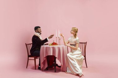 Dönem kıyafetleri giymiş erkek ve kadın romantik bir akşam yemeği yer pembe stüdyo geçmişine rağmen şampanya içerler. 18. yüzyıl yemek gelenekleri. Tarih, retro ve vintage, dönemin karşılaştırması