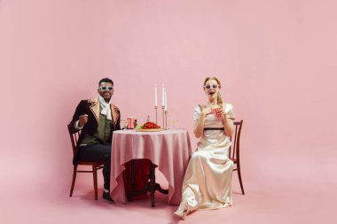 Dönem kostümlü şık Viktorya dönemi çifti masada oturup 3 boyutlu gözlüklerle film izliyor ve pembe stüdyo arka planında patlamış mısırları var. Tarih kavramı, retro ve vintage, çağların karşılaştırması