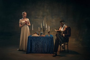 Saten elbiseli kadın, mum ışığında duran şarap kadehini tutarken, adam da eski moda giysiler içinde eski duvar halısı arka planına karşı kitap okuyor. Tarih, mahsul, çağların karşılaştırılması, yeniden yapım konsepti