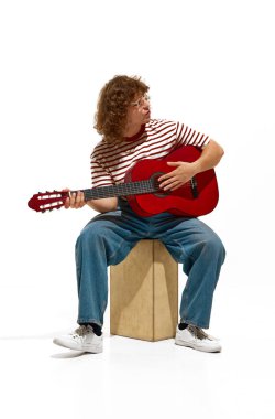 Genç bir adam, kot pantolon giyen kıvırcık saçlı bir gitarist ve çizgili bir gömlek beyaz arka planda izole gitar çalarak akustik bir set sergiliyor. Müzik, festival, konser, hobi, yetenek, performans kavramı