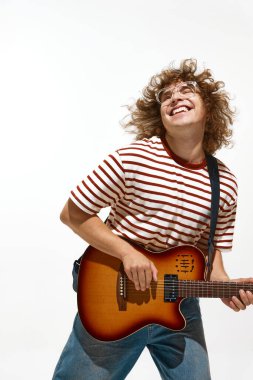 Çizgili gömlekli ve kot pantolonlu enerjik bir gencin portresi beyaz arka planda izole edilmiş duygularla gitar çalıyor. Müzik, festival, konser, hobi, yetenek, performans kavramı