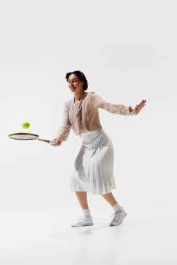 Beyaz arka planda soyutlanmış raketle tenis oynayan etekli ve bluzlu zarif genç bir kadının tam boy dikey görüntüsü. Spor anlayışı, retro ve vintage, aktif yaşam tarzı, hobi