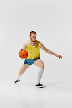 Sarı formalı ve mavi şortlu odaklanmış genç bir adamın tam boy görüntüsü, beyaz arka planda topu olan hareketli bir basketbolcu. Spor, retro ve vintage, aktif yaşam tarzı, hobi konsepti