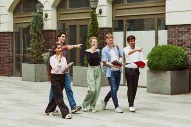 Bir grup çok ırklı insan, erkek ve kız, üniversitenin dışında yürüyen ve heyecan dolu bir yeri işaret eden öğrenciler. Eğitim kavramı, üniversite, çok kültürlü öğrenciler, birlik