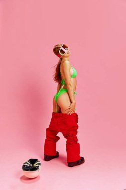 Yeşil bikinili ve kırmızı pantolonlu genç bir kadın pembe stüdyo geçmişine karşı neşeli ve kendinden emin bir poz veriyor. Tatil eğlencesi. Tatil kavramı, turizm, aktif yaşam tarzı, moda, gençlik