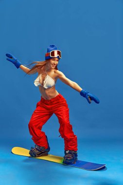 Snowboard Macerası. Bikini üstlü ve kırmızı kar pantolonlu genç bir kadın, mavi stüdyo arka planında parlak bir gülümsemeyle neşeyle snowboard sürüyor. Tatil, turizm, aktif yaşam tarzı, moda kavramı