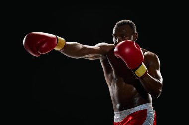 Yumruk at. Afro-Amerikan kaslı bir adam, kırmızı eldivenli ve şortlu bir boksör siyah arka planda izole edilmiş yumruklar eğitiyor. Profesyonel spor anlayışı, aktif yaşam tarzı, vücut, güç