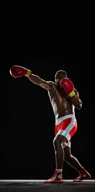 Uzun boylu kaslı bir adam, kırmızı şortlu üstsüz bir boksör ve yumruk eğitimi görmüş bir eldiven. Siyah arka planda izole edilmiş. Spor anlayışı, aktif yaşam tarzı, vücut, güç. Dikey poster