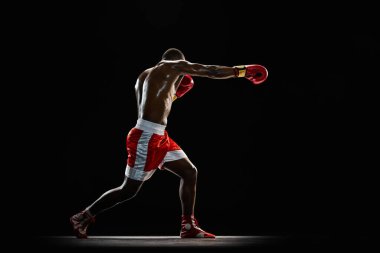 Yumruk at. Kaslı üstsüz bir adam, kırmızı üniformalı bir boksör maçtan önce antrenman yapıyor, siyah arka planda yoğun antrenman yapıyor. Profesyonel spor anlayışı, aktif yaşam tarzı, vücut, güç