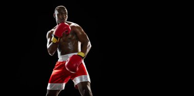 Afrikalı kaslı bir adam, kırmızı eldivenli ve şortlu bir boksör savunma pozisyonunda duruyor, siyah arka planda izole edilmiş bir eyleme hazır. Profesyonel spor anlayışı, aktif yaşam tarzı, vücut, güç