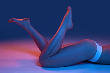 İnce, pürüzsüz kadın bacakları. İç çamaşırlarıyla, mavi stüdyo arkaplanına karşı neon ışıkta poz veren bir model. Kadın güzelliği, vücut ve cilt bakımı, zindelik, sağlık