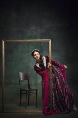 Meraklı, zarif genç bir kadının portresi, kraliyet mensubu, koyu yeşil arka planla ilgilenen bir resim çerçevesine bakıyor. Geleceğin gizemi. Tarih kavramı, çağların karşılaştırması, yeniden yapım