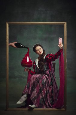 Genç ortaçağ kraliyet ailesinden birinin portresi, altın çerçevenin içinde oturan dev şarap kadehiyle ve cep telefonuyla eski yeşil arka planda selfie çeken bir kadın. Tarih kavramı, restoran