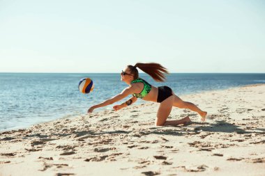 Güneş gözlüklü ve spor giyimli genç bir kadının dinamik görüntüsü. Hareket halinde, aksiyon halinde, oyuncu kumsaldaki voleybol için hamle yapıyor. Spor anlayışı, yaz, doğa, aktif yaşam tarzı, gençlik