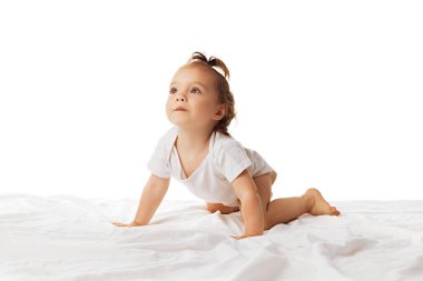 Meraklı bir kız bebek, bebek tulumuyla yatakta sürünüyor, oynuyor, beyaz arka planda tek başına hareket ediyor. Aktif çocuk. Çocukluk kavramı, bakım, sağlık, çocuk, yaşam tarzı, annelik