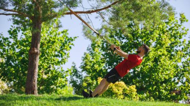 Zengin yeşil parkta TRX egzersizi yapan kaslı bir adam destek için ağaç dallarını kullanıyor ve mükemmel form ve odaklanma gösteriyor. Spor, vücut ve sağlık anlayışı, aktif yaşam tarzı