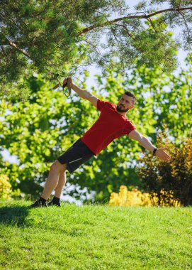 TRX egzersiz oturumu. Yeşil parkın dışında spor yaparken kaslı, sakallı, kırmızı tişörtlü bir adam. Egzersiz için traks kayışları kullanıyor. Spor, vücut ve sağlık anlayışı, aktif yaşam tarzı