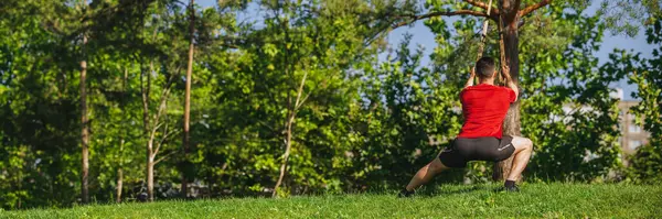 Arkadan bak. Spor giyiminde sporcu adam dışarıda antrenman yapıyor, yeşillik parkında spor yapıyor, çömelme egzersizleri yapıyor. Spor anlayışı, vücut ve sağlık hizmetleri, aktif yaşam tarzı. Pankart