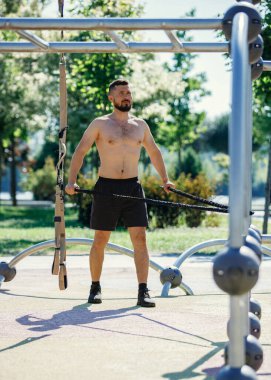 Açık hava parkında direniş bantlarıyla egzersiz yapan, siyah şort giyen, antrenmanına odaklanan üstsüz bir adam. Kaslı vücut şekli. Spor, vücut ve sağlık anlayışı, aktif yaşam tarzı, dayanıklılık