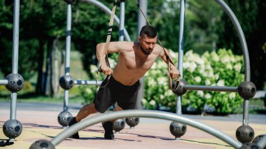 Parkta sporcu, üstsüz antrenman yapıyor, metal çerçeveye bağlı süspansiyon kayışlarıyla egzersiz yapıyor. TRX eğitim oturumu. Şınav. Spor, vücut ve sağlık anlayışı, aktif yaşam tarzı, dayanıklılık