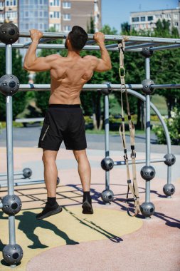 Açık hava fitness parkındaki metal bir çubukta siyah şort giymiş üstsüz bir adam barfiks çekiyor. Sabah antrenmanı. Spor, vücut ve sağlık anlayışı, aktif yaşam tarzı, dayanıklılık