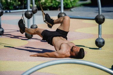 Açık hava fitness parkında yerde yatan gömleksiz bir adam, süspansiyon kayışlarıyla bacak kaldırma, açık hava egzersizi yapıyor. Spor, vücut ve sağlık anlayışı, aktif yaşam tarzı, dayanıklılık