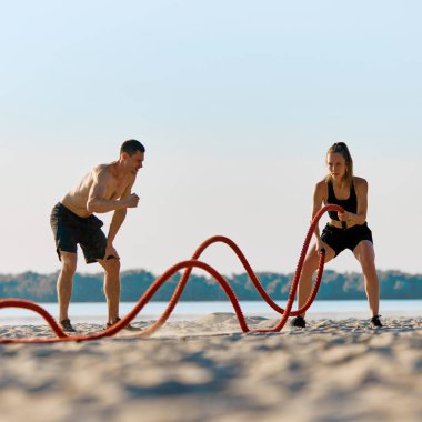 Siyah şortlu gömleksiz adam dışarıda kumsalda savaş halatlarıyla egzersiz yapan atletik kadını motive ediyor. Spor anlayışı, aktif ve sağlıklı yaşam tarzı, vücut bakımı