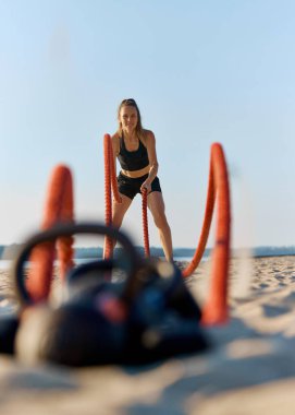 Siyah spor elbiseli atletik bir kadın kumsalda kırmızı savaş halatları kullanıyor, açık mavi gökyüzünün altında gölde. Yoğun spor eğitimi. Spor anlayışı, aktif ve sağlıklı yaşam tarzı, dayanıklılık