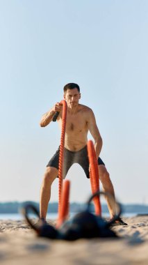 Siyah şortlu odaklanmış bir sporcu, plajda kırmızı savaş halatlarıyla egzersiz yapan üstsüz bir adam, güç ve dayanıklılık gösteriyor. Spor anlayışı, aktif ve sağlıklı yaşam tarzı. Dikey resim