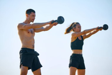 Siyah spor kıyafetleri içinde üstsüz bir adam ve kadın açık havada kettlebell salıncakları yapıyor, açık bir arka planda antrenman yapıyorlar. Spor anlayışı, aktif ve sağlıklı yaşam tarzı, vücut bakımı, fitness