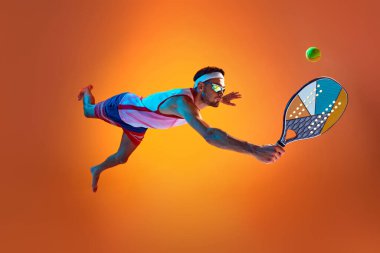 Spor giyim ve güneş gözlüklü genç bir adam neon ışıkta turuncu stüdyo arka planına karşı dinamik bir şekilde plaj tenisi oynuyor. Spor anlayışı, aktif yaşam tarzı, hobi, oyunlar, rekabet