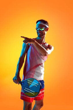 Spor giyimli ve güneş gözlüklü bir adam Padel tenis raketi tutuyor, neon ışıkta turuncu arka plana bakıp gülümsüyor. Spor anlayışı, aktif ve sağlıklı yaşam tarzı, hobi, oyunlar, rekabet