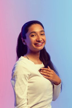 Beyaz gömlekli, uzun siyah saçlı, pembe ve mavi arkaplanda neon ışıkta duran neşeli bir şekilde gülümseyen, neşe ve mutluluğu ifade eden genç bir kadın. İnsan duyguları ve gençlik kavramı