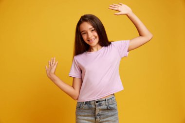 Selamlar. Mutlu, gülümseyen, sıradan giyinmiş, neşeli poz veren ve sarı stüdyo geçmişine el sallayan güzel bir genç kız. Pozitif titreşimler. İnsan duyguları kavramı, çocukluk, gençlik, eğitim