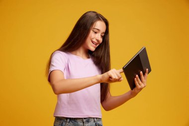 Kahverengi uzun saçlı, pembe tişörtlü mutlu bir genç kız, sarı stüdyo geçmişine karşı eğlence ve eğitim için tablet kullanıyor. İnsan duyguları kavramı, çocukluk, gençlik, eğitim