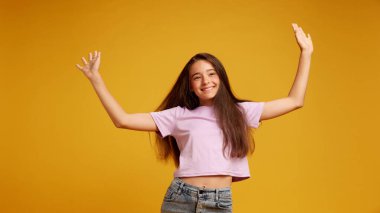 Mutlu, pembe tişörtlü, neşeli bir şekilde dans eden ve sarı stüdyo geçmişine karşı sevinçle poz veren genç kızın portresi. İnsan duyguları kavramı, çocukluk, gençlik, eğitim