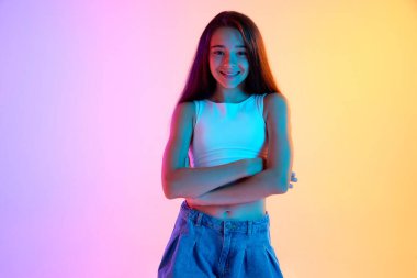 Portrait of happy teen girl in casual clothes, white top and jeans, posing with smile against gradient pink yellow background in neon light. Concept of human emotions, childhood, youth, education clipart