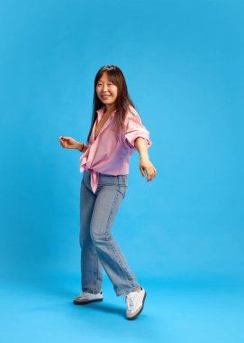 Kot pantolonlu, pembe gömlekli, mavi stüdyo arka planına poz veren kendine güvenen Asyalı genç bir kadının tam boy dikey görüntüsü. İnsan duyguları, gençlik, yaşam tarzı, gündelik moda kavramı