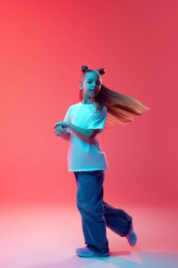 Gündelik giysiler içinde, beyaz tişört ve kot pantolon içinde pembe arka planda dans eden genç bir kız resmi. Çocukluk kavramı, insan duyguları, moda