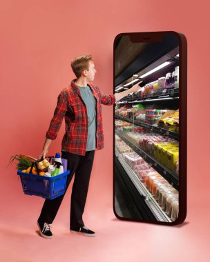 Kırmızı ekose gömlekli bir adam internetten alışveriş yapıyor, elinde alışveriş sepeti tutuyor, büyük akıllı telefon ekranlarına dokunuyor ve pembe arka planda market raflarını gösteriyor. Çevrimiçi alışveriş, yemek, teslimat uygulaması kavramı